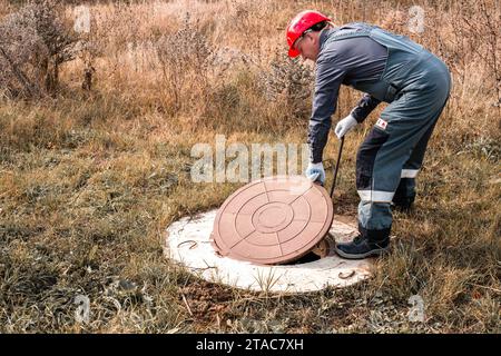 Ein Arbeiter mit einem Schutzhelm hebt einen Mannlochdeckel auf einen septischen Brunnen. Inspektion und Instandhaltung von Kanalisationssystemen im ländlichen Raum. Stockfoto