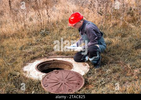 Ein Versorgungsarbeiter in einem Helm zeichnet die Messwerte eines Wasserzählers in einem Betonbrunnen auf. Inspektion und Wartung von Leitungen. Stockfoto