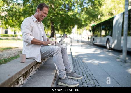 Freiberuflicher Programmierer mittleren Alters, der auf einer Bank sitzt und online am Laptop im Stadtpark arbeitet. Stockfoto