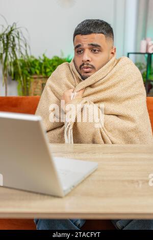 Kranker, kranker indischer Mann, der an einem Laptop arbeitet, der in Karo gewickelt ist, sitzt zitternd vor kaltem trinken heißem Kaffee im unbeheizten Heimbüro, ohne Schulden zu heizen. Ungesunder Araber versuchen, sich aufzuwärmen Stockfoto