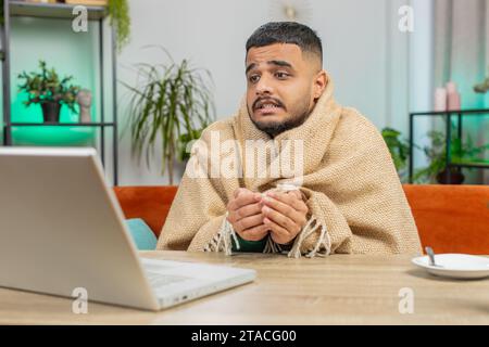 Kranker, kranker indischer Mann, der an einem Laptop arbeitet, der in Karo gewickelt ist, sitzt zitternd vor kaltem trinken heißem Kaffee im unbeheizten Heimbüro, ohne Schulden zu heizen. Ungesunder Araber versuchen, sich aufzuwärmen Stockfoto