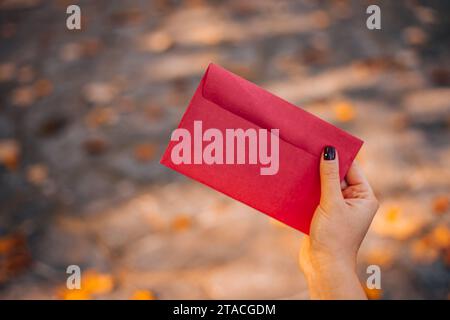 Eine Frau öffnet einen roten Umschlag, während sie auf einer Bank im Stadtpark sitzt. Roter Umschlag ohne Inschrift auf Herbstlaub-Hintergrund Stockfoto