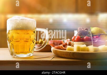 Bierkrug auf Holzbank mit Snacks daneben und rustikalem Küchenhintergrund. Vorderansicht. Stockfoto