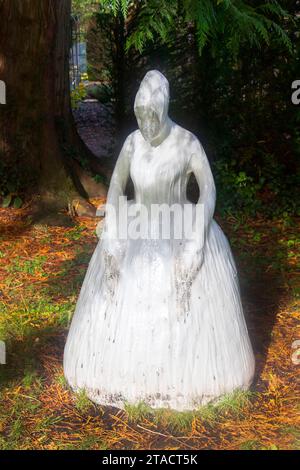 Eine weiße geisterhafte Statue einer Kapuzenfigur in einem Wald, umgeben von Grün und hohen Bäumen, die Schatten auf den Boden werfen. Stockfoto