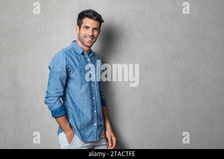 Glücklicher, lächelnder Mann, der sich an die graue Wand lehnt. Porträt eines stolzen Mittelmannes, isoliert auf grauem Hintergrund. Junger, lässiger hispanischer Mann gegen graue Mauerschauerin Stockfoto