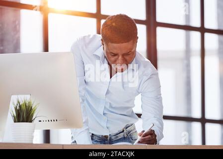 Seriöser, aufmerksamer Geschäftsmann der Millennials, der am Tisch steht und sich Notizen in einem Notizbuch macht Stockfoto