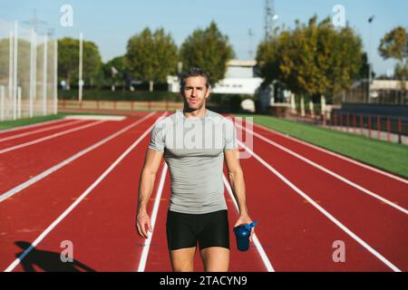 Reifer Fitness-Mann auf roter Laufbahn, läuft und sieht blaue Flasche in der Hand. In grauem T-Shirt und schwarzer Hose gekleidet. Vorderansicht. Kopierbereich. Stockfoto