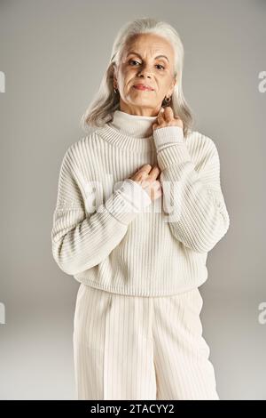 Attraktive und grauhaarige Frau mittleren Alters in weißem Winteroutfit mit grauem Hintergrund Stockfoto