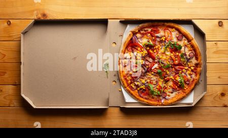 Peperoni-Pizza mit Paprika, Zuckermais und Rucola in offener Schachtel auf natürlichem Holzküchentisch, flacher Lay View mit Kopierraum Stockfoto