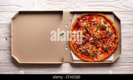 Pepperoni Pizza mit Paprika, Zuckermais und Rucola in offener Schachtel auf weißem Holzküchentisch mit flachem Blick. Kopierbereich Stockfoto