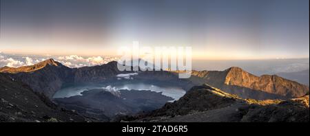 Panoramablick auf einen vulkanischen Kratersee bei Sonnenaufgang, umgeben von zerklüfteten Bergen und Wolken, die lange Schatten über die dramatische Landschaft werfen. Stockfoto