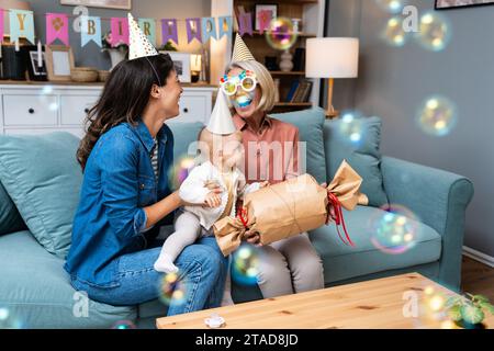 Mehrgenerationenfamilie mit Geschenken auf einer Indoor-Geburtstagsfeier. Tochter mit Baby, die mit lustigen Hüten den Geburtstag der Mutter oder Großmutter feiert Stockfoto