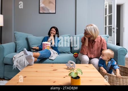 Junge unverantwortliche Mutter schaut fern und isst Popcorn, während ihre Mutter, Großmutter, sich um das Baby kümmert. Sorglose Mutter achtet nicht auf ihr Chi Stockfoto