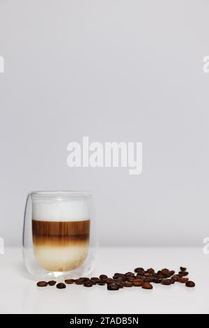 Nahaufnahme einer doppelten Glas Tasse frischen Latte Coffee mit Kaffeebohnen auf weißem Tisch und Kopierraum Stockfoto