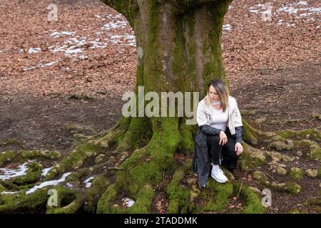 Frau, die auf moosbedeckten Baumwurzeln in einem Wald sitzt, ein weißes Hemd, eine schwarze Hose und weiße Schuhe trägt Stockfoto