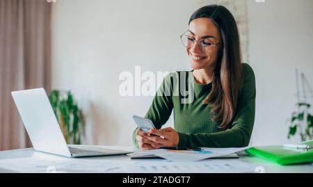 Nahaufnahme eines Porträts einer selbstbewussten jungen charmanten Geschäftsfrau, die ihr Smartphone nutzt, während sie zu Hause mit einem Laptop arbeitet Stockfoto