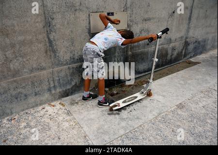 Ein kleiner Junge schlürft erfrischendes Wasser und hält seinen Roller im Parc des Chuamettes an einem modernen Trinkbrunnen fest. Stockfoto