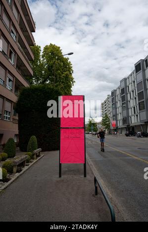 Ein lebhaftes pinkfarbenes Poster kündigt eine Museumsausstellung in Genf an, während ein Mann auf seinem Roller vorbeigleitet und das Stadtleben mit Kunst verbindet. Stockfoto