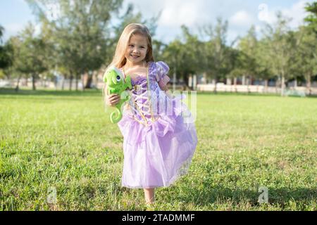 Kleines blondes Mädchen mit langen Haaren, das Rapunzel mit einem Chamäleon Pascal verkörpert. Blondes kleines Mädchen in einem lila Kleid. Halloween Kostüm, Halloween Stockfoto