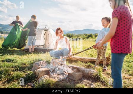 Drei Schwestern machen Picknick, rösten Marshmallows und Süßigkeiten auf Stöcken über Lagerfeuer, während zwei Brüder das grüne Zelt aufbauten. Glückliche Familie, Outd Stockfoto