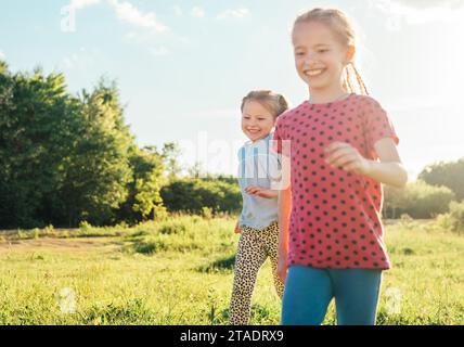 Zwei kleine Schwestern laufen und lachen auf der sommerlich hohen grünen Wiese mit einem hellen Sonnenuntergang Licht Hintergrund. Sorglose Kindheit, Familienwerte Stockfoto