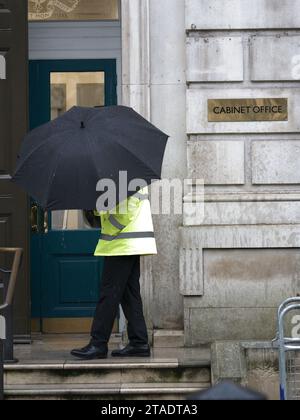 Sicherheitsbeamter mit Schirm an einem Eingang zum Kabinettsgebäude der britischen Regierung in Whitehall, London, England, während eines regnerischen Tages Stockfoto