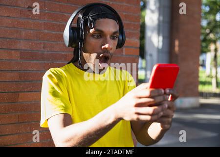 Junger schwarzer Mann in der Stadt mit einem durag, der sehr überrascht ist, was er auf seinem Handy sieht Stockfoto