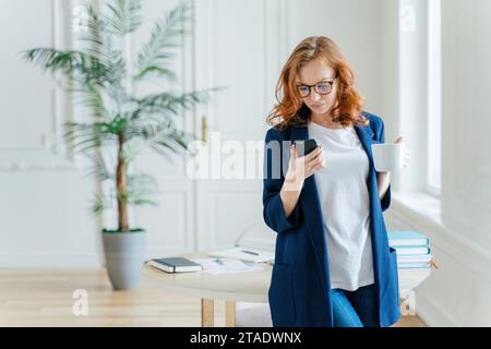 Fokussierte Frau in blauem Blazer mit Smartphone und Kaffeetasse in einem Heimbüro Stockfoto