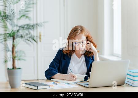 Konzentrierte rothaarige Geschäftsfrau, die in einem hellen Büro an einem Laptop mit Notizen arbeitet Stockfoto