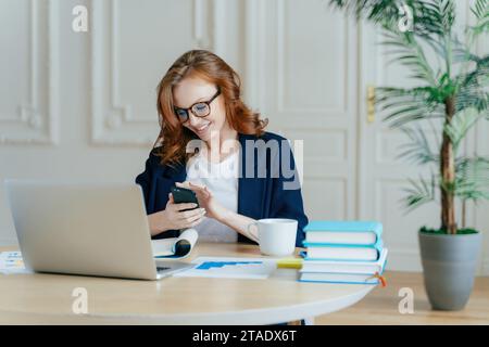 Geschäftsfrau lächelt ihr Smartphone im Büro mit Laptop und Kaffee an, produktiver Tag Stockfoto