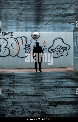 Silhouette einer Person, die in einer dunklen Fußgängerunterführung läuft, mit Graffiti an der Wand hinter sich. Stockfoto