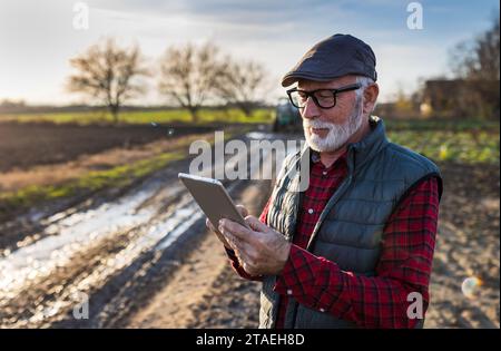 Porträt eines Seniorenbauers mit Brille und Kappe, der im Winter auf dem Feld steht und vor dem Traktor auf einem Tablet arbeitet Stockfoto