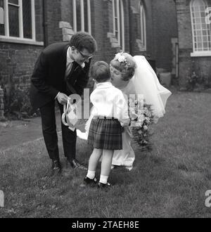 1960er Jahre, historisch, Hochzeit, auf dem Gelände einer Kirche, ein Kuss auf die Wange von einer Braut in ihrem Hochzeitskleid für ein kleines Pageboy im Kilt, nachdem sie ihr das traditionelle Geschenk des Glücklichen Pferdeschmuts überreicht hat, England, Großbritannien. Stockfoto