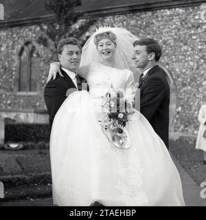 1960er Jahre, historisch, vor der Kirche bei einer Hochzeit, eine Braut in ihrem Hochzeitskleid und Schleier wird vom Bräutigam und besten Mann getragen, England, Großbritannien. Stockfoto