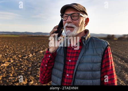 Porträt eines Seniorenbauers, der im Winter auf dem Feld steht und mit dem Handy spricht Stockfoto