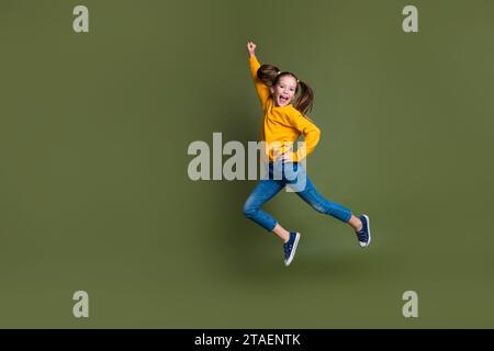 Foto in voller Länge von überfröhlichem Kind mit Pferdeschwänzchen gekleidetes gelbes Hemd, das die Faust hochspringt, isoliert auf khakifarbenem Hintergrund Stockfoto