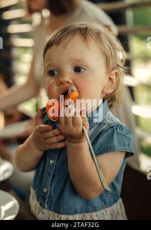 Porträt eines kleinen Mädchens, das eine Aprikose mit einer Gabel isst. Vertikaler Rahmen. Stockfoto