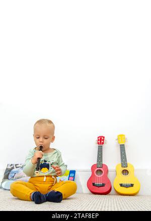 Ein in Gelb und Grün gekleidetes Kleinkind setzt sich hin und pfeift neben zwei Ukulelen Stockfoto