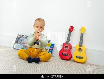 Ein in Gelb und Grün gekleidetes Kleinkind setzt sich hin und pfeift neben zwei Ukulelen Stockfoto