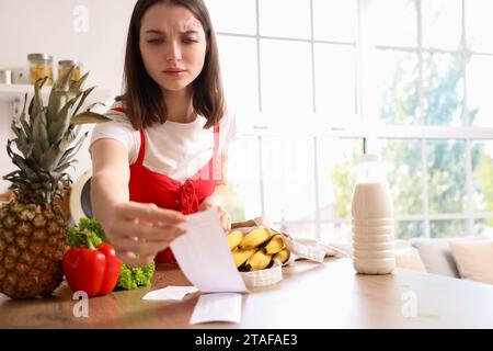 Verärgerte junge Frau mit Ladenquittung und Essen in der Küche. Preissteigerungskonzept Stockfoto