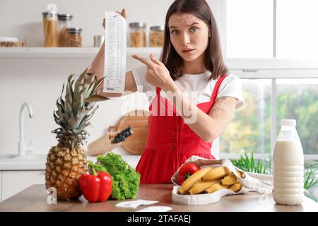 Eine junge Frau mit Essen, die auf den Kaufbeleg in der Küche zeigt. Preissteigerungskonzept Stockfoto