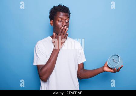 Ein junger Erwachsener afroamerikanischer Ethnizität schaut sich die Zeit an und deckt den Mund unter Schock. Schwarze männliche Person, die immer pünktlich ist, eine Wanduhr trägt und ängstlich erscheint. Stockfoto