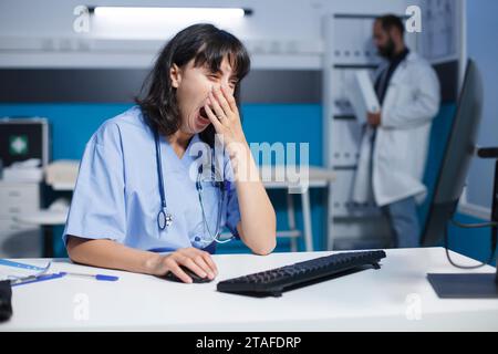 Erschöpfte Ärztin in einem gut beleuchteten Krankenhausraum am Schreibtisch mit dem Desktop-pc. Das Bild zeigt eine gähnende kaukasische Krankenschwester, die an einem Computer in einer Arztpraxis arbeitet. Stockfoto