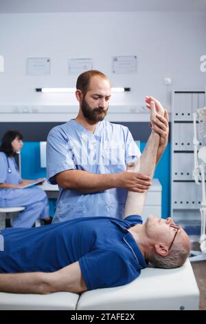 Männlicher älterer Patient, der durch einen Arzt osteopathisch wegen Armschmerzen versorgt wird. Kaukasischer pensionierter alter Mann mit Hilfe einer Krankenschwester in Blauschutt durch physiotherapeutische Übung. Stockfoto