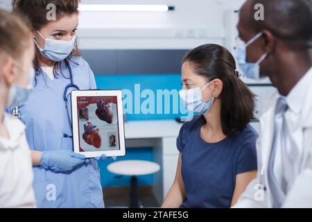 Detaillierte Abbildung von medizinischem Fachpersonal, das weibliche Patienten mit einer Tablette über das menschliche Herz erklärt. Die Krankenschwester hält ein Gerät, während der männliche Arzt die Wichtigkeit der medizinischen Untersuchungen beschreibt. Stockfoto