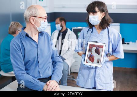 Die Krankenschwester in blauen Peelings erklärt die kardiologische Behandlung, die auf dem Bildschirm des Geräts angezeigt wird. Arzt und älterer Patient mit Gesichtsmasken analysieren das Bild des Herzens auf einem digitalen Tablet. Frau mit Gadget. Stockfoto