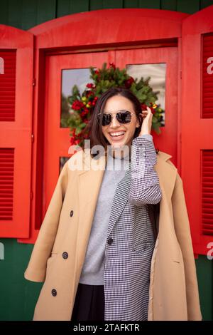 Fröhliches Lächeln kommt aus dem Weihnachtsgeschäft. Frau lächelt im Wintermantel mit Sonnenbrille steht in der Nähe des Weihnachtsgeschäfts. Frau, die auf der Straße steht, im Wintermantel Sonnenbrille, die Haare repariert. Vertikal Stockfoto