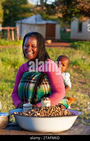 Eine afrikanische Straßenverkäuferin mit Zöpfen trägt ein Kind im Rücken, das in eine Decke gewickelt ist Stockfoto