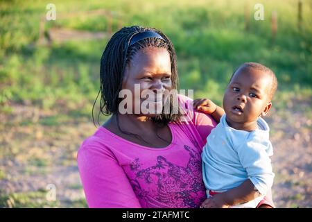 Eine afrikanische Dorfmutter mit Zöpfen trägt ein Kind in ihren Armen Stockfoto