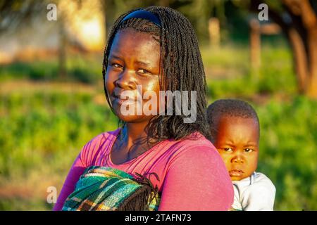 afrikanische Dorfmutter mit Zöpfen trägt das Kind in eine Decke gewickelt im Rücken Stockfoto